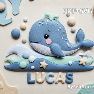 Pode incluir: Uma baleia de feltro azul claro com bochechas rosa e detalhes brancos, repousando sobre ondas de feltro em tons de azul e verde. As ondas têm bolhas e estrelas de feltro. O nome "LUCAS" está escrito em letras de feltro. O texto "PDF+SVG" é visível.