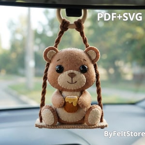 May include: A felt hanging ornament featuring a brown bear sitting on a swing, holding a honey pot. The bear has black eyes and a light tan belly. The text PDF+SVG and ByFeltStore are visible. The ornament hangs from a wooden ring.