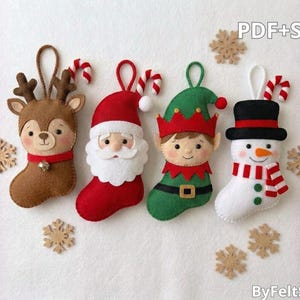 May include: Four felt Christmas ornaments shaped like stockings featuring a brown reindeer, Santa Claus, an elf, and a snowman. Each ornament has a hanging loop and a small candy cane accent. Six wooden snowflake cutouts surround the figures.