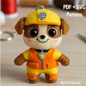 May include: A felt dog keychain in yellow, orange, brown, and cream colors. The dog is wearing a yellow helmet with a badge and an orange vest. The text "PDF + SVG Pattern" is visible in the upper right corner.