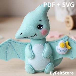 May include: A light blue felt pterodactyl with stitched details, rosy cheeks, and a smiling face. The wings are spread, and it holds a small egg and star. Text on the image reads "PDF + SVG" and "ByFeltStore".