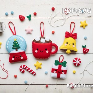 Pode incluir: Enfeites de Natal de feltro feitos à mão, incluindo um globo de neve, uma caneca de chocolate quente, um sino e uma caixa de presente. Os enfeites são vermelhos, brancos, amarelos e azuis, com rostos sorridentes e detalhes decorativos. O texto na imagem diz "PDF+SVG" e "ByFeltStore".