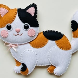Pode incluir: Um gato de feltro feito à mão em branco, preto e laranja. O gato tem olhos grandes pretos, bochechas rosadas e um laço rosa. O desenho é costurado com linha branca. O gato está em pé.