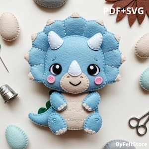 May include: A light blue felt triceratops dinosaur with white horns, a beige belly, and pink cheeks. The dinosaur has large black eyes and a smiling mouth. The image also includes the text "PDF+SVG" and "ByFeltStore".