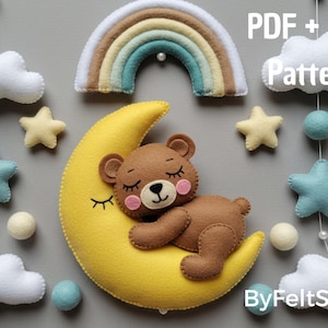 Può includere: Mobile per bambini in feltro con un orsetto marrone addormentato su una luna crescente gialla, circondato da stelle, nuvole e un arcobaleno. Il mobile include il testo "PDF + SVG Pattern" e "ByFeltStore".