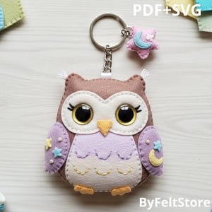May include: A handmade felt owl keychain with a silver keyring and a small star charm. The owl is brown, lavender, and cream-colored, with embroidered details and large, expressive eyes. The text "PDF+SVG" and "ByFeltStore" are visible.