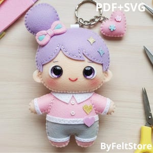Pode incluir: Um chaveiro de boneca de feltro feito à mão com um coque e laço de lavanda, usando uma camisa rosa e shorts cinza. A boneca tem olhos grandes e um rosto sorridente. Um pequeno pingente em forma de coração rosa está anexado. O texto "PDF+SVG" é visível.