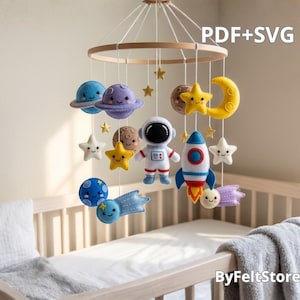 Op de afbeelding: Een baby mobile van vilt met planeten, sterren, een raket, een astronaut en een maan. De mobile hangt aan een houten ring boven een wieg. De tekst "PDF+SVG" is zichtbaar.
