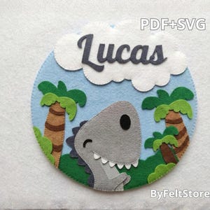 Pode incluir: Decoração de parede circular em feltro com um dinossauro cinzento, palmeiras e o nome "Lucas" em escrita cinzenta escura sobre um céu azul com nuvens brancas. Os textos "PDF+SVG" e "ByFeltStore" também são visíveis.