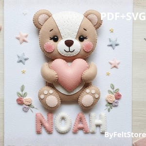 Pode incluir: Um urso de feltro feito à mão segurando um coração rosa, com o nome "NOAH" abaixo. O urso é marrom claro com uma mancha branca na cabeça e detalhes em rosa. Estrelas e flores decorativas circundam o urso. O texto "PDF+SVG" e "ByFeltStore" também são visíveis.