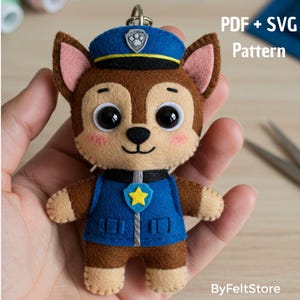 Op de afbeelding: Een handgemaakte vilten sleutelhanger met een cartoonhond in een blauw politie-uniform en -pet. De hond heeft bruine vacht, roze oren en grote zwarte ogen. De tekst "PDF + SVG Pattern" en "ByFeltStore" zijn zichtbaar.