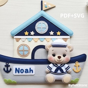 Puede incluir: Decoración de fieltro con forma de barco con techo azul marino, paredes beige y casco gris. Un oso de peluche con un traje de marinero está sentado en el barco. El nombre "Noah" está en el barco. El texto "PDF+SVG" y "ByFeltStore" también son visibles.