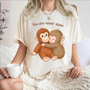 May include: Cream-colored t-shirt featuring a cartoon illustration of two hugging monkeys. The text "You are never alone" is printed above the monkeys. The shirt is made of a soft material.