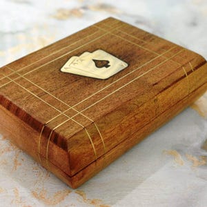 Puede incluir: Una caja rectangular de madera con un diseño de naipes. La caja es de madera marrón claro con líneas incrustadas de color dorado. Una pequeña incrustación de color marfil presenta dos naipes, incluido el As de Picas.