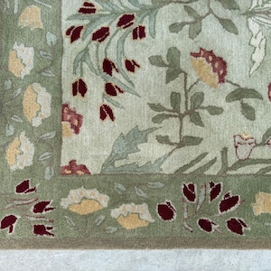 Puede incluir: Primer plano de una sección de una alfombra con un diseño floral. La alfombra presenta un fondo verde claro con un borde verde oscuro. El diseño incluye flores color crema, acentos burdeos y hojas verdes. Ideal para el hogar.