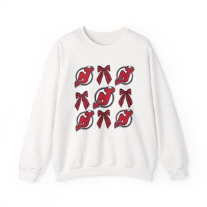 May include: White sweatshirt with a repeating pattern of red bows and the New Jersey Devils logo. The logo is a stylized 'NJ' with a hockey stick and a devil's tail. The design is repeated across the front.