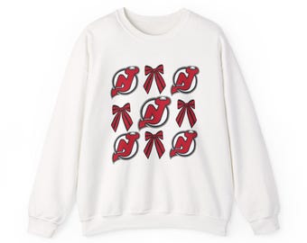 May include: White sweatshirt featuring a pattern of red bows and the New Jersey Devils logo. The logo is a stylized 'NJ' with a hockey stick and a devil's tail. The design is repeated across the front of the sweatshirt.