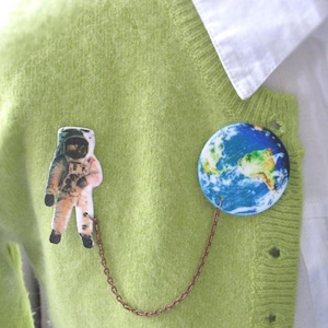 Könnte beinhalten: Ein skurriles Astronauten- und Erd-Anstecknadel-Set. Der Astronauten-Pin ist in einem Raumanzug, und der Erd-Pin zeigt eine detaillierte Karte. Die Pins sind durch eine zarte Kette verbunden und an einem hellgrünen Pullover befestigt.
