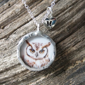 May include: Silver-toned owl pendant necklace. The pendant features a detailed owl illustration within a circular frame. A small heart charm with the letter "M" hangs near the clasp. The necklace is displayed on a textured wooden surface.