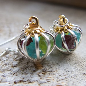 Two cages hold crystal gemstones attached to ear wires to make personalized earrings. Birthstones fill the sterling silver cages with gold top. Sold by What A Novel on Etsy.