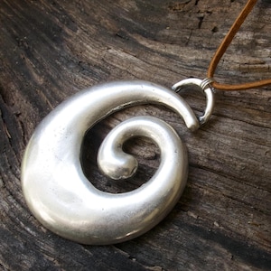 May include: A silver-toned pendant necklace featuring a large, swirling spiral design. The pendant is suspended from a brown leather cord. The necklace is displayed on a rustic wooden surface, highlighting the pendant's metallic sheen.