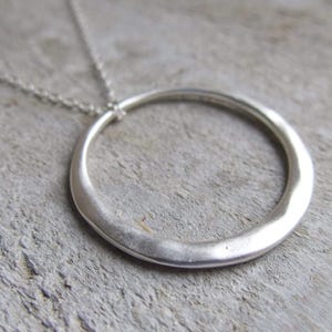 May include: A silver pendant necklace with a large, uneven circular pendant. The pendant is suspended from a fine chain. The necklace has a matte finish and a minimalist design, suitable for everyday wear.