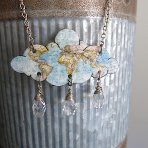 Op de afbeelding: Zilveren ketting met een wolk-vormige hanger. De hanger heeft een wereldkaart ontwerp in blauw, geel en roze. Drie heldere kristallen druppels hangen aan de wolk. De ketting is geplaatst tegen een getextureerde, metalen achtergrond.