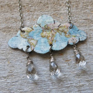 May include: A silver chain necklace featuring a cloud pendant with a world map design. The cloud is light blue with a map print in shades of tan and brown. Three clear, faceted crystal teardrops dangle below the cloud.