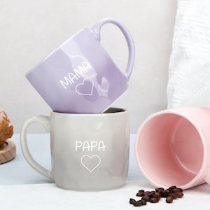 Puede incluir: Tres tazas de cerámica en tonos lavanda, gris y rosa. La taza lavanda tiene la palabra "MAMA" y un corazón, la taza gris tiene "PAPA" y un corazón. Granos de café esparcidos cerca de la taza rosa.