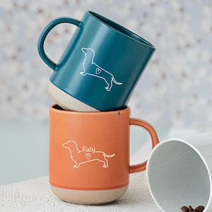 May include: Stacked ceramic mugs in teal and orange, each featuring a white outline of a dachshund dog with a heart. The orange mug has the name "Ruby" above the heart. The mugs have a speckled beige base and handle.