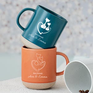 May include: Two ceramic mugs, one teal and one orange, stacked. Each mug has a white cat design in a heart, with "best friends" and two names printed below. The mugs have a speckled, light-coloured base. The mugs are for beverages.