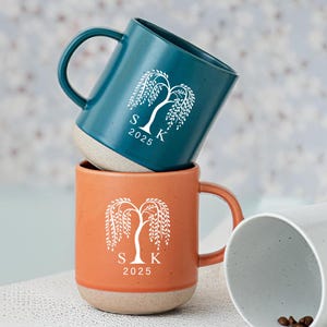 Puede incluir: Dos tazas de cerámica, una verde azulado y otra naranja, apiladas. Cada taza presenta un diseño de sauce llorón blanco con las iniciales "S K" y el año "2025". Las tazas tienen una base moteada de color claro y un asa curva.