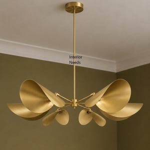 May include: A brass chandelier featuring six curved, petal-shaped shades arranged around a central stem. The fixture has a circular ceiling canopy and a thin vertical rod. The text Interior Needs is visible on the central stem of the light.