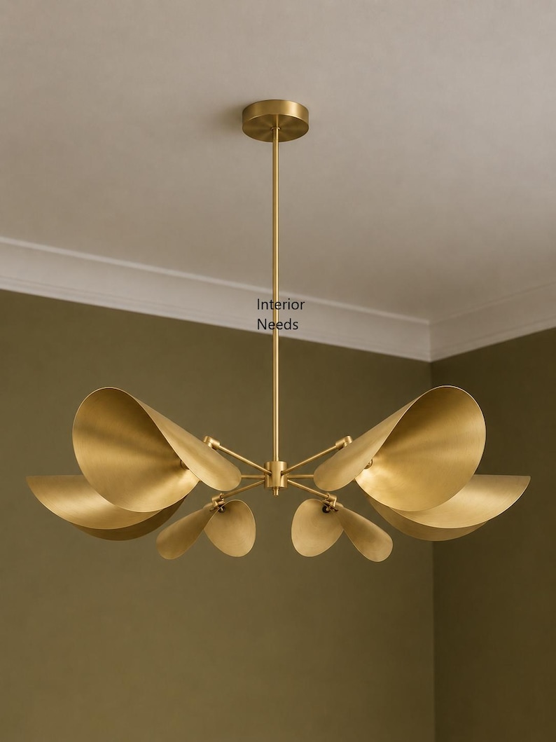 May include: A brass chandelier featuring six curved, petal-shaped shades arranged around a central stem. The fixture has a circular ceiling canopy and a thin vertical rod. The text Interior Needs is visible on the central stem of the light.