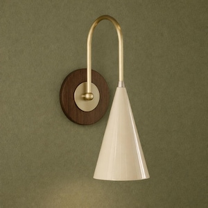 Peut inclure: Une applique murale composée d'une plaque arrière en bois foncé circulaire avec un disque de montage central en laiton. Un bras courbé en laiton s'élève puis redescend pour soutenir un abat-jour conique lisse de couleur crème.