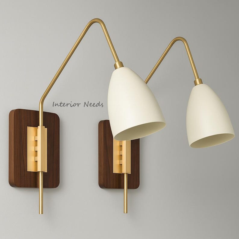 May include: Two wall sconces with a mid-century modern design. Each has a dark wood rectangular base, a gold-toned arm, and an off-white, cone-shaped shade. The text "Interior Needs" is visible on the wall.