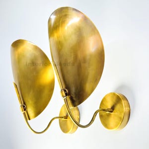May include: A pair of brass wall sconces with a unique design. Each sconce features a curved arm connected to a circular base and supports a large, leaf-shaped shade. The sconces are made of polished brass and are mounted on a white wall.