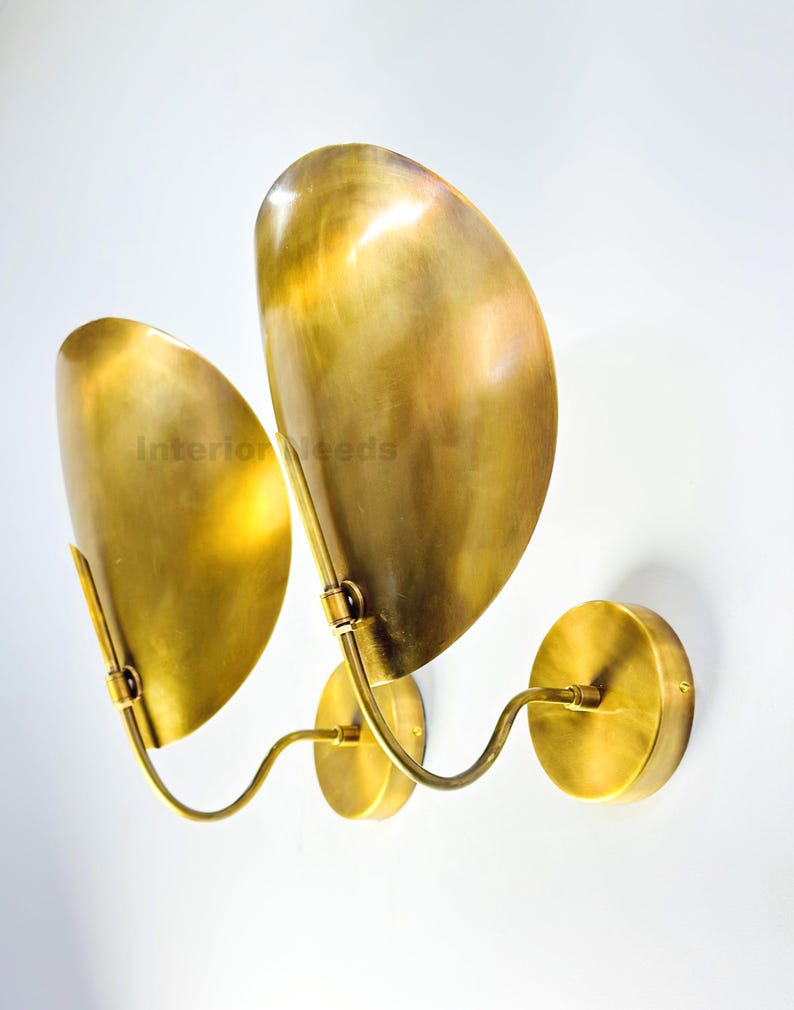 May include: A pair of brass wall sconces with a unique design. Each sconce features a curved arm connected to a circular base and supports a large, leaf-shaped shade. The sconces are made of polished brass and are mounted on a white wall.