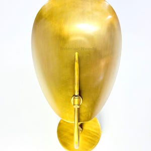 May include: A gold-colored wall sconce with a rounded, elongated shade and a circular base. The sconce has a vertical rod and a small, circular mounting plate. The text "Interior Needs" is visible on the shade.
