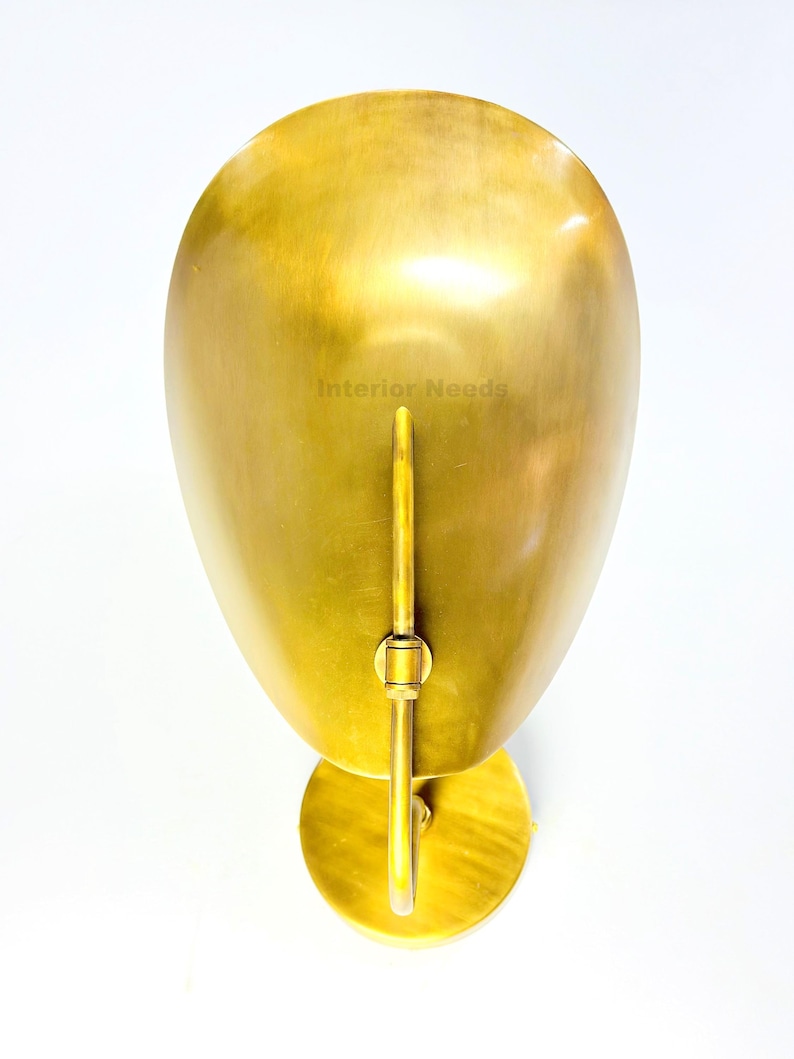 May include: A gold-colored wall sconce with a rounded, elongated shade and a circular base. The sconce has a vertical rod and a small, circular mounting plate. The text "Interior Needs" is visible on the shade.