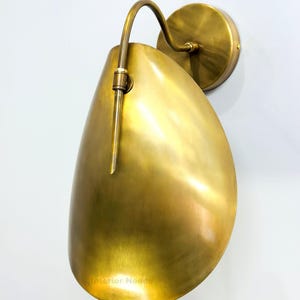 May include: A brass wall sconce with a teardrop-shaped shade and curved arm. The sconce has a circular base and a warm, golden finish. The design is minimalist and modern, suitable for interior lighting.