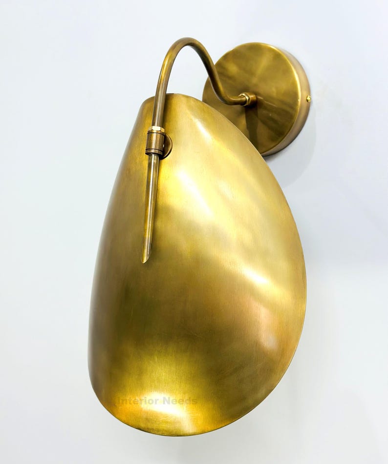 May include: A brass wall sconce with a teardrop-shaped shade and curved arm. The sconce has a circular base and a warm, golden finish. The design is minimalist and modern, suitable for interior lighting.