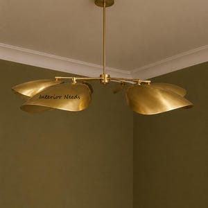 May include: A brass chandelier featuring a central rod and four curved, petal-shaped shades extending outward. The metal has a polished gold-toned finish. The text Interior Needs is printed on one of the shades. The fixture hangs from a circular ceiling mount.