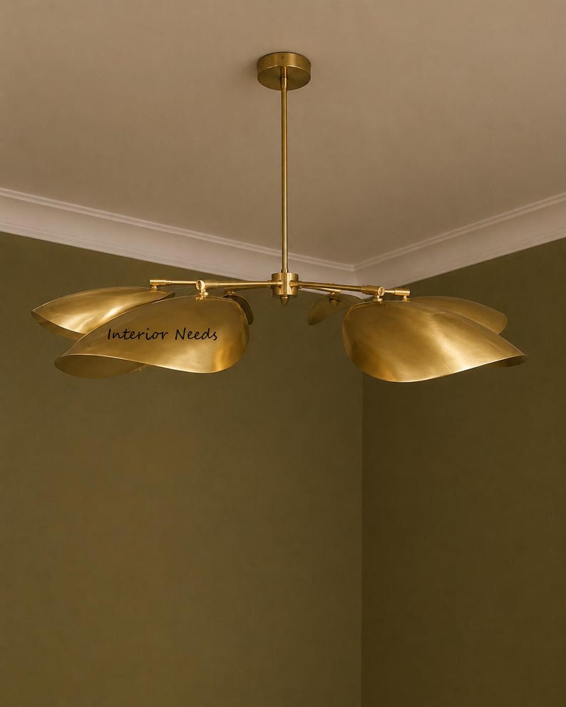 May include: A brass chandelier featuring a central rod and four curved, petal-shaped shades extending outward. The metal has a polished gold-toned finish. The text Interior Needs is printed on one of the shades. The fixture hangs from a circular ceiling mount.