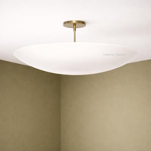 May include: A modern, white, oval-shaped ceiling light fixture with a brass-colored stem and ceiling mount. The fixture is suspended from the ceiling and has the text "Interior Needs" on the side.