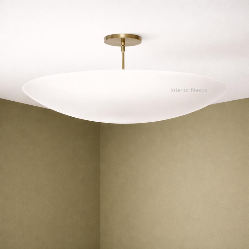 May include: A modern, white, oval-shaped ceiling light fixture with a brass-colored stem and ceiling mount. The fixture is suspended from the ceiling and has the text "Interior Needs" on the side.