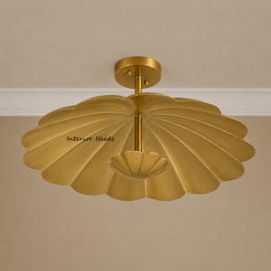 May include: A gold-toned metal flush mount ceiling light fixture featuring a large, scalloped, shell-shaped shade and a smaller, matching scalloped decorative element at the center. The text Interior Needs is printed on the shade surface.