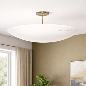 May include: A modern, white, oval-shaped ceiling light fixture with a brass-colored stem and ceiling mount. The fixture is suspended from the ceiling and has the text "Interior Needs" on the side. The light is a soft, diffused glow.