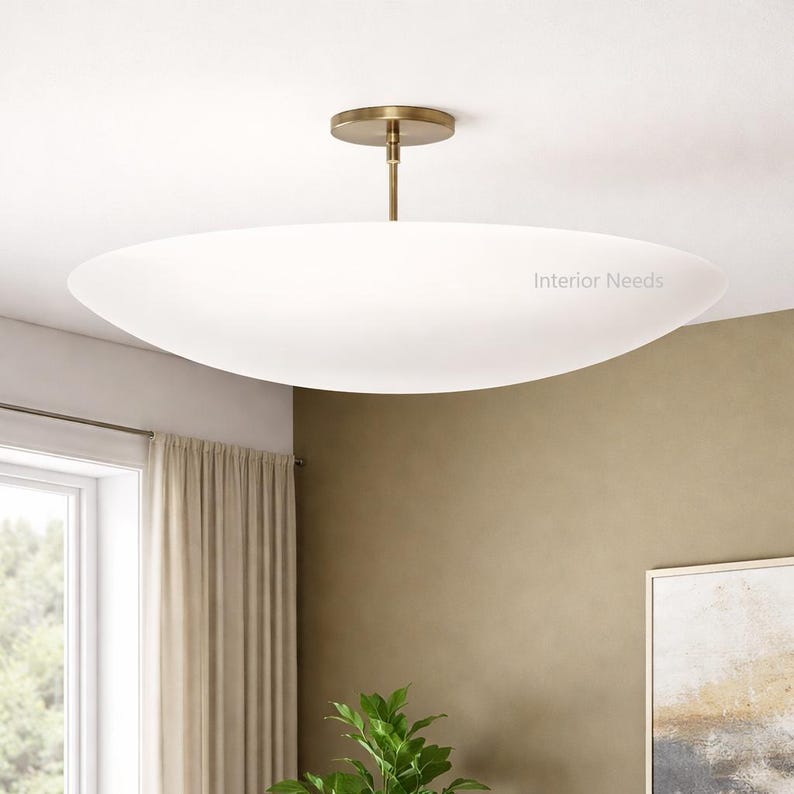 May include: A modern, white, oval-shaped ceiling light fixture with a brass-colored stem and ceiling mount. The fixture is suspended from the ceiling and has the text "Interior Needs" on the side. The light is a soft, diffused glow.