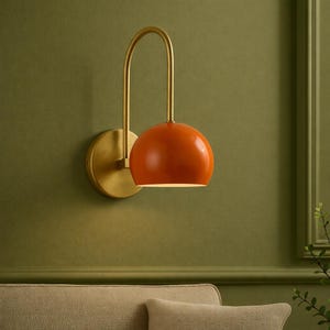 May include: A wall-mounted lamp featuring a bright orange dome-shaped shade attached to a curved brass arm. The fixture includes a circular brass wall plate that connects the arm to the wall, providing a downward-facing light source.